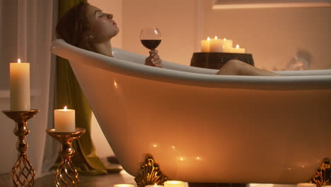 Schönes-Mädchen-In-Einer-Badewanne-Mit-Kerzen-In-Gläsern-Auf-Dem-Boden-Und-Flauschigen-Federn-Auf-Einem-Weißen-Tisch