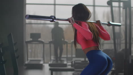 The-young-woman-in-pink-top-and-blue-leggins-is-preparing-herself-for-important-competitions.-She-is-doing-squats-with-an-empty-bar-with-a-right-technique-in-gym-at-the-morning-workout.