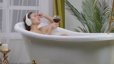 Eine-Junge,-Schöne,-Kaukasische-Brünette-Liegt-Im-Badezimmer-Und-Hört-Musik-In-Weißen-Kopfhörern,-Erholt-Sich-Vom-Stress-Und-Trinkt-Entspannt-Rotwein-Aus-Dem-Glas