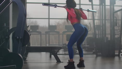 The-girl-in-pink-top-and-blue-leggins-is-doing-squats-with-an-empty-bar.-She-is-holding-the-bar-behind-her-neck-on-shoulders.