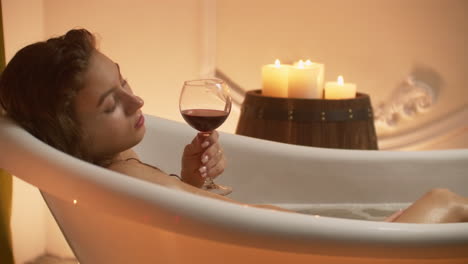 Woman-bathing-with-pleasure-lying-down-in-the-tub-with-foam-and-drinking-red-wine-spending-time-in-luxury-spa-resort
