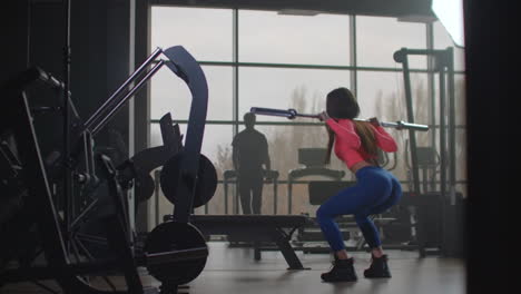 The-sportswoman-in-pink-top-and-blue-leggins-is-preparing-herself-for-important-competitions.-She-is-doing-squats-with-an-empty-bar-with-a-right-technique.