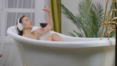 Pretty-young-woman-listening-to-music-in-headphones-while-taking-bath-and-drink-red-wine-from-a-glass.-Attractive-girl-relaxing-has-rest-at-home.-Bathroom-eco-friendly-interior.-Stress-relief-and-feel-good.-Nice-content-enjoyment