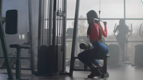 A-young-girl-in-blue-leggings-and-a-pink-top-performs-exercises-on-a-group-of-back-muscles.-Training-in-a-room-with-large-Windows-in-the-background-a-woman-uses-an-ellipsoid-and-a-treadmill.