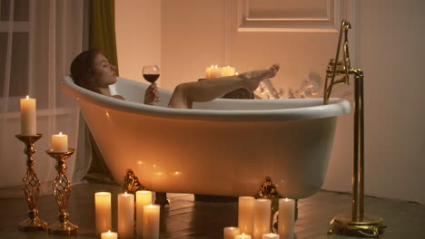 young-sexy-woman-with-straight-naked-body-and-clean-legs-lying-and-relaxing-in-white-foam-bath-tub-with-candles-around-in-light-bathroom-drink-alcohol-from-wine-glass-indoors