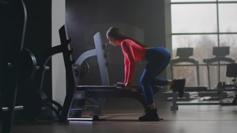 The-woman-in-short-pink-top-is-working-on-her-back-in-the-gym.-She-leans-on-a-bench-with-her-right-part-of-a-body-and-training-her-left-hand-and-left-part-of-the-back.