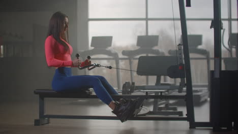 The-woman-with-a-strong-and-slim-body-in-blue-leggins-and-pink-top-performs-an-exercise-on-the-muscles-of-the-back-and-shoulder-blades-in-gym.-Productive-workout-for-a-healthy-body.