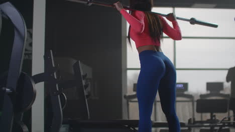 The-bodybuilder-woman-in-pink-top-and-blue-leggins-is-preparing-herself-for-important-competitions.-She-is-doing-squats-with-an-empty-bar-with-a-right-technique.