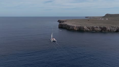 Un-Solo-Barco-Catamarán-Navega-Frente-A-La-Costa-De-Pont-Den-Gil,-Menorca,-España-Al-Mediodía-Con-La-Vela-Arriba