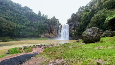 Hunua-Waterfall-flows-into-muddy-river-on-rainy-day,-wide-shot-in-slow-motion