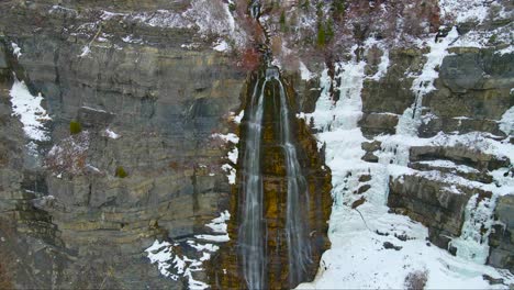 Epic-drone-scenic-waterfall-over-frozen-ice,-rocks,-cliffs,-and-snow-on-Mount-Timpanogos-in-American-Fork,-Utah-while-snowflakes-fall-in-foreground
