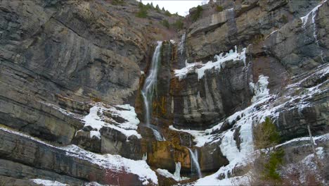 Epischer-Drohnen-szenischer-Wasserfall-über-Gefrorenem-Eis,-Felsen,-Klippen-Und-Schnee-Auf-Dem-Mount-Timpanogos-In-American-Fork,-Utah,-Während-Schneeflocken-Im-Vordergrund-Fallen