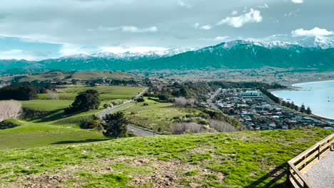 Rolling-grassy-hills-and-coastal-town-with-snowy-mountains,-New-Zealand