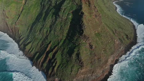 Strong-ocean-waves-crash-on-cliff-base-of-Ponta-Vigia,-drone-tilt-up-to-Ponta-do-Pargo-lighthouse,-Portugal