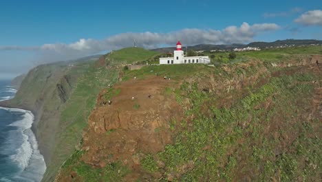 Orbit-around-tourists-exploring-sheer-cliffs-below-white-lighthouse-in-Madeira-Portugal