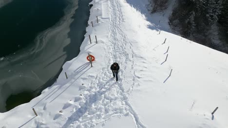 With-head-bowed-down,-a-man-is-walking-on-a-snowy-path-beside-the-frozen-lake,-located-on-top-of-a-mountain-at-Engelberg,-Brunni,-in-Bahnen,-Switzerland