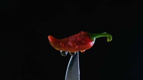 Red-wet-Jalapeno-pepper-on-a-black-background-pinched-from-bottom-by-a-silver-knife-with-shots-of-flashing-light-coming-from-lower-left-side