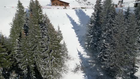 Approaching-drone-shot-revealing-a-log-cabin-opposite-a-pond-on-a-snowy-slope-where-cable-cars-stop-by-on-their-way-to-the-top-of-the-mountain,-in-Engelberg,-Brunni,-in-Bahnen,-Switzerland