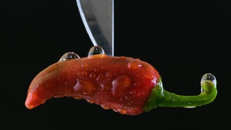 Red-wet-Jalapeno-pepper-pinched-by-a-silver-knife-slowly-dropping-from-top-on-a-black-background