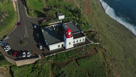 Drone-ascend-tilt-down-to-red-and-white-lighthouse-building-on-sheer-rocky-cliff-at-midday