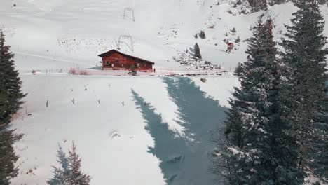 Zipline-drone-shot-of-a-log-cabin-beside-a-frozen-pond-and-right-next-to-a-snowy-mountain-slope,-located-at-Engelberg,-Brunni,-in-Bahnen,-Switzerland