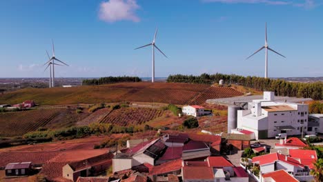 Drone-flying-forward-over-village-in-Torres-Vedras,-Portugal,-with-wind-turbines-rotating-while-revealing-the-coastline-on-the-background