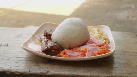 Woman-puts-on-the-table-a-plate-with-banku,-a-typical-Ghanaian-dumpling,-accompanied-by-fish-and-other-garnishes,-Ghanaian-lunch