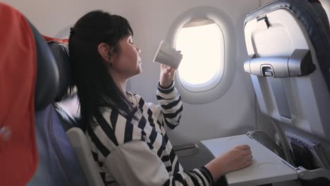 Woman-sitting-inside-airplane-and-looking-at-window-and-drinks-coffee.