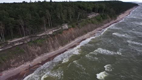 Unveiling-a-towering-coastal-cliff-along-the-Baltic-Sea-in-Lithuania,-captured-by-a-drone,-showcasing-turbulent-waves-and-vibrant-green-waters-with-atmospheric-hues