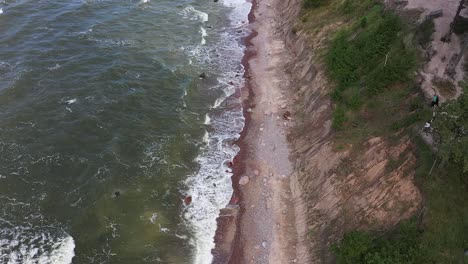 Overhead-drone-views-showcase-the-rugged-charm-of-a-high-cliff-coast-in-Lithuania's-Baltic-Sea,-where-powerful-waves-meet-the-enchanting-green-sea,-painted-with-moody-colors