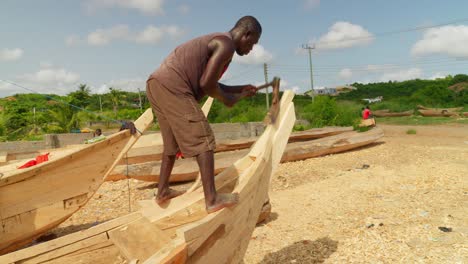 African-man-carving-bow-of-wooden-boat-with-adze-in-village-shipyard