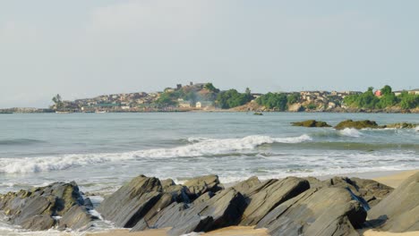 Waves-hitting-sandy-beach-with-black-rocks,-coastal-town-beyond-bay