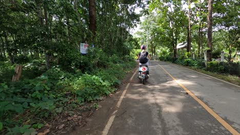 Young-female-traveler-riding-a-scooter-on-a-serene-road-surrounded-by-lush-greenery-in-Pai,-Thailand