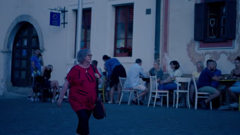 Mujer-Blanca-Mayor-Con-Pelo-Gris,-Gafas-Y-Camiseta-Roja-Caminando-Por-La-Histórica-Ciudad-De-Bardejov-En-Eslovaquia-Durante-Un-Atardecer-De-Hora-Azul-En-Cámara-Lenta,-Rodeada-De-Gente