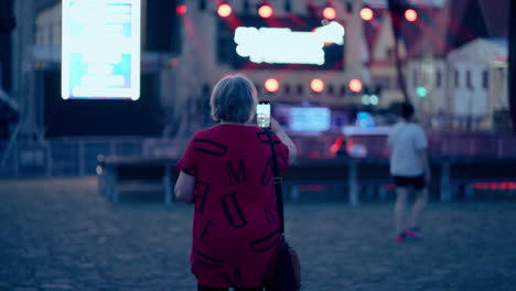 Older-Caucasian-Woman-making-Video-with-her-Smartphone-of-a-Stage-with-flickering-red-and-white-Lights-during-a-blue-hour-at-night-in-an-old-historical-town-Bardejov-in-Slovakia-in-slow-motion