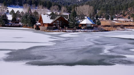 First-snow-ice-frozen-covered-white-Evergreen-Lake-House-Rocky-Mountain-landscape-scene-morning-front-range-Denver-aerial-cinematic-drone-Christmas-ice-skating-hockey-blue-sky-backwards-pan-up-motion