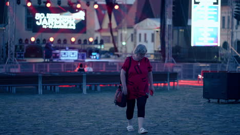 Older-caucasian-Woman-wearing-Glasses-and-red-T-shirt-walking-away-from-Stage-with-flickering-red-and-white-Lights-and-smiling-in-slow-motion-in-an-old-historical-City-Bardejov-in-Slovakia