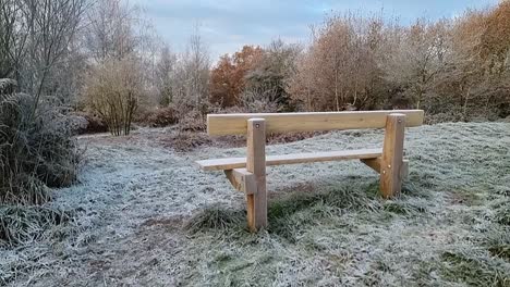 Empty-wooden-park-bench-on-frosty-ice-coated-grassy-hillside-overlooking-autumn-woodland-foliage
