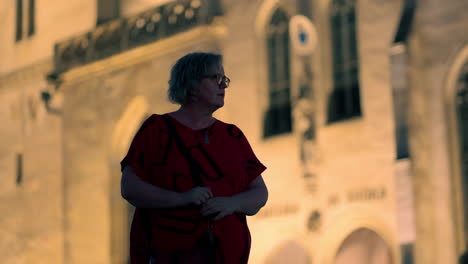 Older-white-Woman-with-glasses-standing-and-looking-around-in-front-of-illuminated-historical-European-church-at-night-in-slow-motion