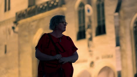 Older-white-Woman-with-red-T-shirt-and-glasses-standing-and-looking-around-in-front-of-illuminated-historical-European-church-at-night-in-slow-motion