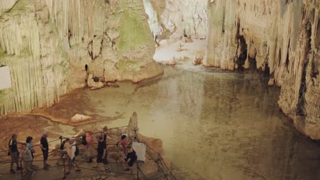 Several-tourists-walk-with-a-tour-guide-through-Neptune's-grotto,-with-stalactites-and-next-to-an-underground-lagoon
