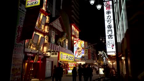 Silhouette-Of-People-Along-Street-In-Shinsekai-Area-Illuminated-By-Neon-Signs-At-Night-In-Osaka