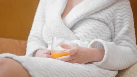 Gorgeous-woman-in-white-bathrobe-relaxing-on-couch-with-glass-of-juice