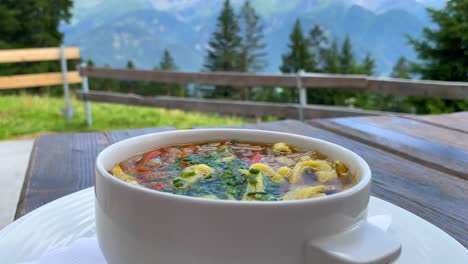 Traditional-Austrian-Frittatensuppe-with-a-beautiful-summertime-mountain-view-in-Tyrol-Austria,-delicious-hot-comforting-soup-in-Serfaus-Fiss-Ladis,-4K-shot