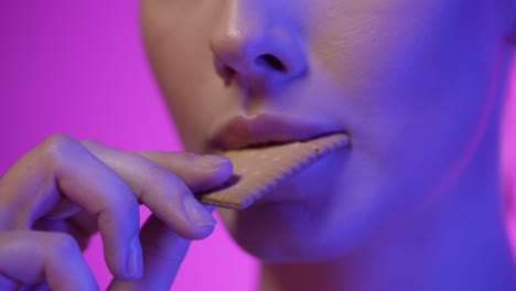Close-up-shot-of-a-young-pretty-woman-eating-a-delicious-cookie-and-enjoying-it-in-front-of-purple-background-in-slow-motion