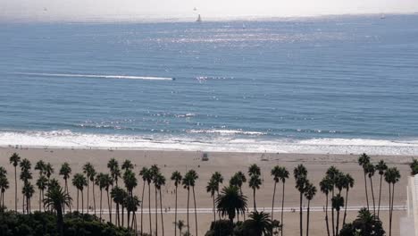 Aerial-View-Of-Santa-Monica-Beach-Lined-With-Palm-Trees-With-Speedboat-Sailing-Across-In-Ocean