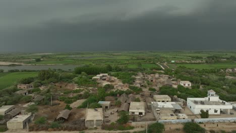 Aerial-drone-rotating-shot-over-rural-village-houses-with-green-farmlands-in-the-background-in-Mirpurkhas,-Sindh,-Pakistan-with-dark-rain-clouds-at-daytime