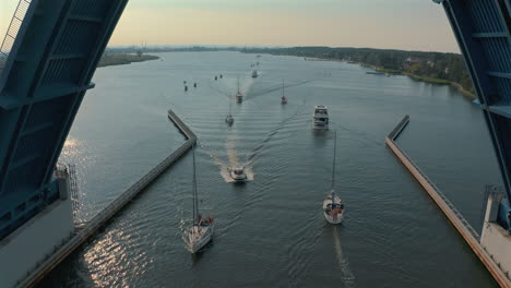Drone-flying-through-opening-bridge-in-Sobieszewo,-Gdańsk-with-passing-boats-at-the-sunset