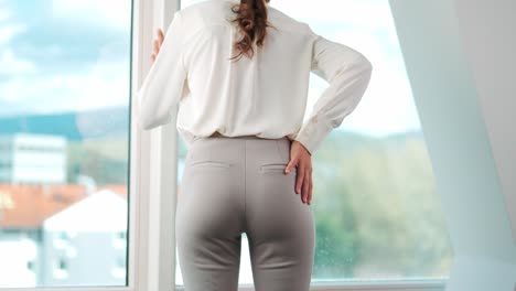Back-view-of-young-woman-with-back-pain-in-front-of-window-in-modern-apartment-office,-back-problems,-incorrect-posture