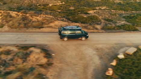 Old-nostalgic-van-bus-with-surfboards-on-roof-drives-along-dusty-road-in-Mediterranean-landscape,-drone-tracking